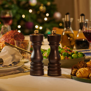 A festive dinner table features the Cole & Mason London Chocolate Wood Mill Christmas Gift Hamper at its center, with food, drinks, and a glowing decorated Christmas tree in the background—ideal for holiday gift giving.