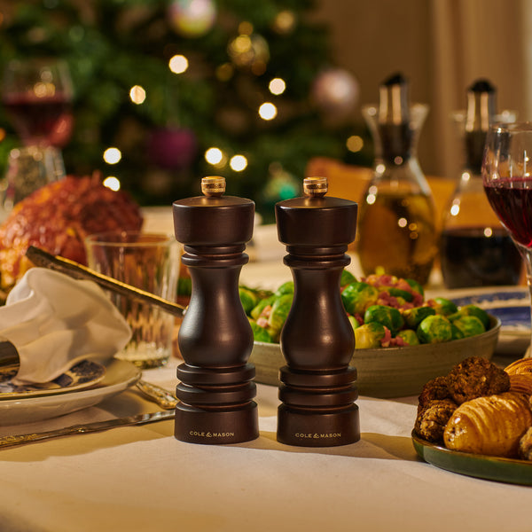 A festive dinner table features the Cole & Mason London Chocolate Wood Mill Christmas Gift Hamper at its center, with food, drinks, and a glowing decorated Christmas tree in the background—ideal for holiday gift giving.