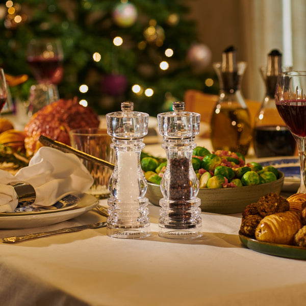 Two clear Cole & Mason grinders from the London Acrylic Mill Christmas Gift Hamper sit on a festive dining table with food and wine, while a glowing Christmas tree in the background adds to the warm holiday atmosphere.