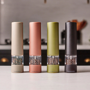 Four Cole & Mason Brixton Electric Mill Stone Set grinders—each in beige, pink, green, and black—stand side by side on a kitchen counter, each filled with peppercorns and equipped with a durable ceramic grinding system. A discreet USB-C charging port is visible on each mill, perfectly complementing the modern kitchen setting against a softly blurred background.