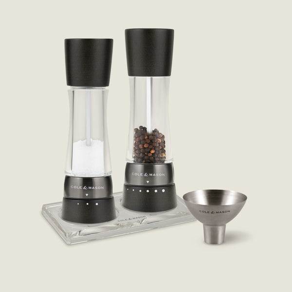 #blackwood&irongrey The Cole & Mason Derwent Salt & Pepper Mill, Acrylic Tray & Funnel Set features two mills with black tops—one for salt, one for peppercorns—displayed on a glass tray beside a stainless steel funnel.