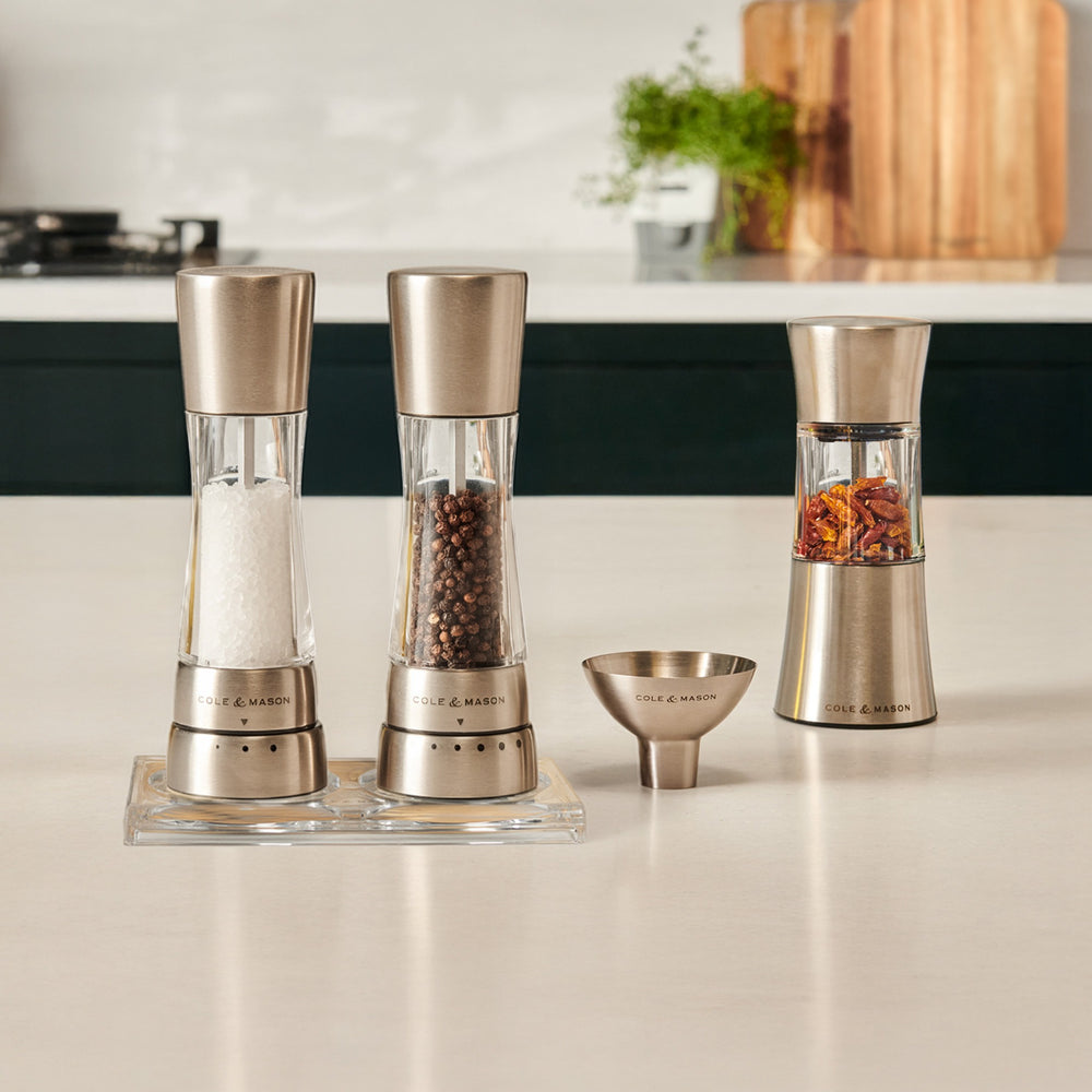 The Cole & Mason Derwent Mill and Stadhampton Chilli Mill Seasoning Set includes matching mills with a funnel and tray, filled with salt, peppercorns, and chili. Both grinders are displayed on a neutral background.