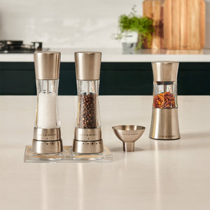 A Cole & Mason Derwent Mill and Stadhampton Chilli Mill Seasoning Set sits on the counter, holding salt, peppercorns, and red chili flakes. A matching funnel and a cutting board are also visible in the background.