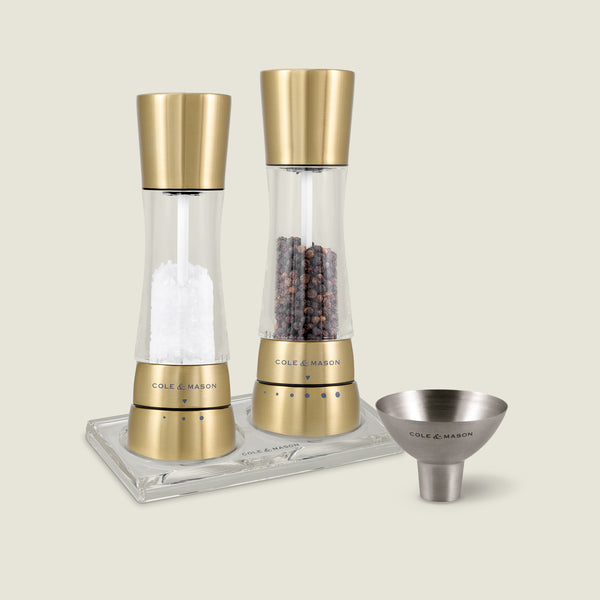#gold The Cole & Mason Derwent Mill Set features gold and clear mills on an acrylic tray, with a stainless steel funnel beside them, all arranged on a light beige background.