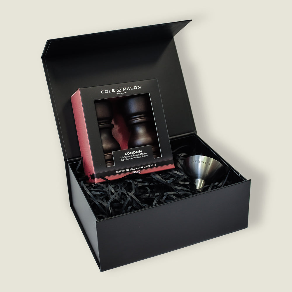 A black gift box with shredded paper holds the Cole & Mason London Chocolate Wood Mill & Shaker Christmas Gift Hamper, featuring wooden mills and shaker, a stainless steel funnel, all neatly arranged inside with the lid open.