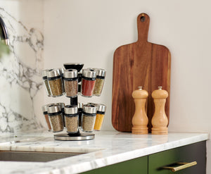 A spice rack with glass jars, a wooden cutting board, and two wooden salt and pepper grinders sit on a marble kitchen countertop near a sink.