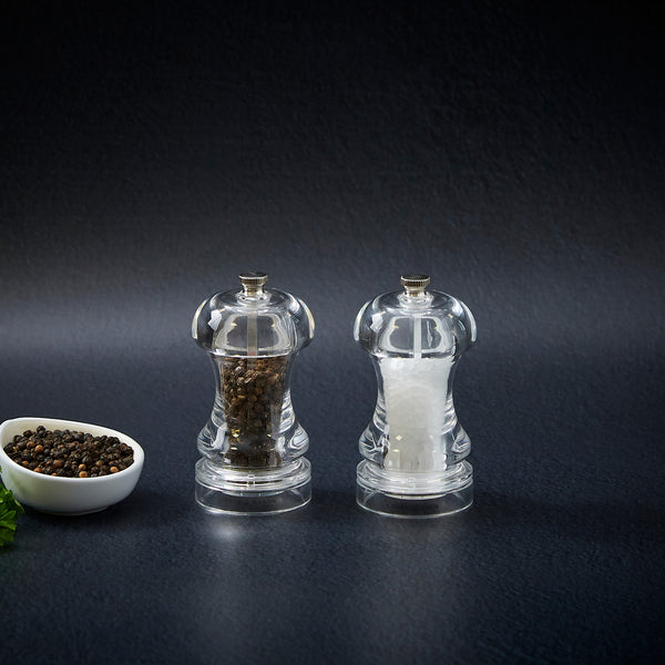 Two clear Cole & Mason Acrylic Capstan Mills—one with black peppercorns, one with white salt—stand side by side on a dark surface. A small bowl of peppercorns completes this perfect Acrylic Capstan Mill Christmas Gift Hamper.