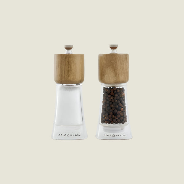 The Cole & Mason Woodbury Salt & Pepper Mill Set features two clear mills with wooden tops, one for salt and one for pepper. Both mills provide adjustable coarseness, all set against a light background for rustic appeal.