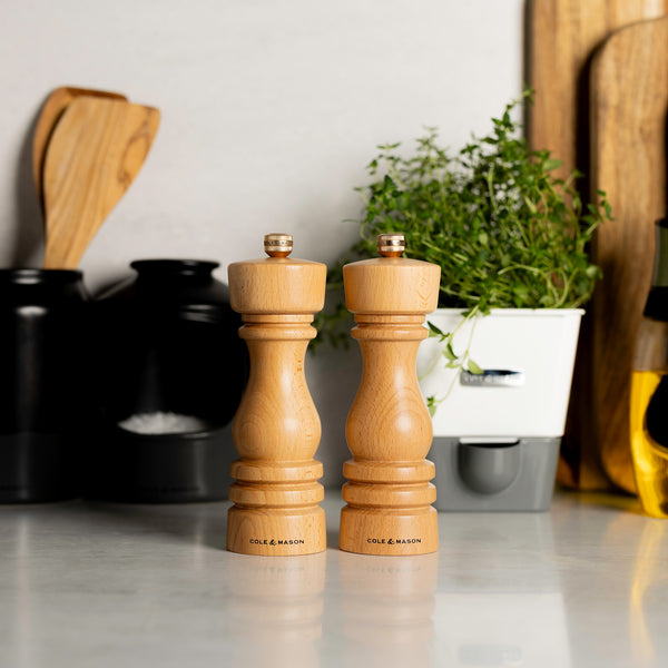 The Cole & Mason London Salt & Pepper Mill Set 180mm Bundle is displayed side by side on a kitchen counter, with potted herbs, olive oil, and utensils visible in the background.