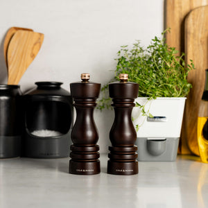 The Cole & Mason London Salt & Pepper Mill Set 180mm Bundle sits on the kitchen counter, alongside a black salt cellar, wooden utensils, fresh herbs in a white planter, and a bottle of oil in the background.
