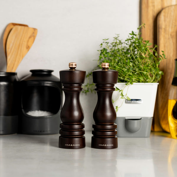 The Cole & Mason London Salt & Pepper Mill Set 180mm Bundle sits on the kitchen counter, alongside a black salt cellar, wooden utensils, fresh herbs in a white planter, and a bottle of oil in the background.