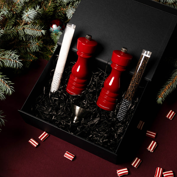 The Cole & Mason London Red Gloss Mill Christmas Gift Hamper features a salt & pepper mill set, metal funnel, and seasoning tubes on black shredded paper, all set in a festive scene with pine branches and peppermint candies.