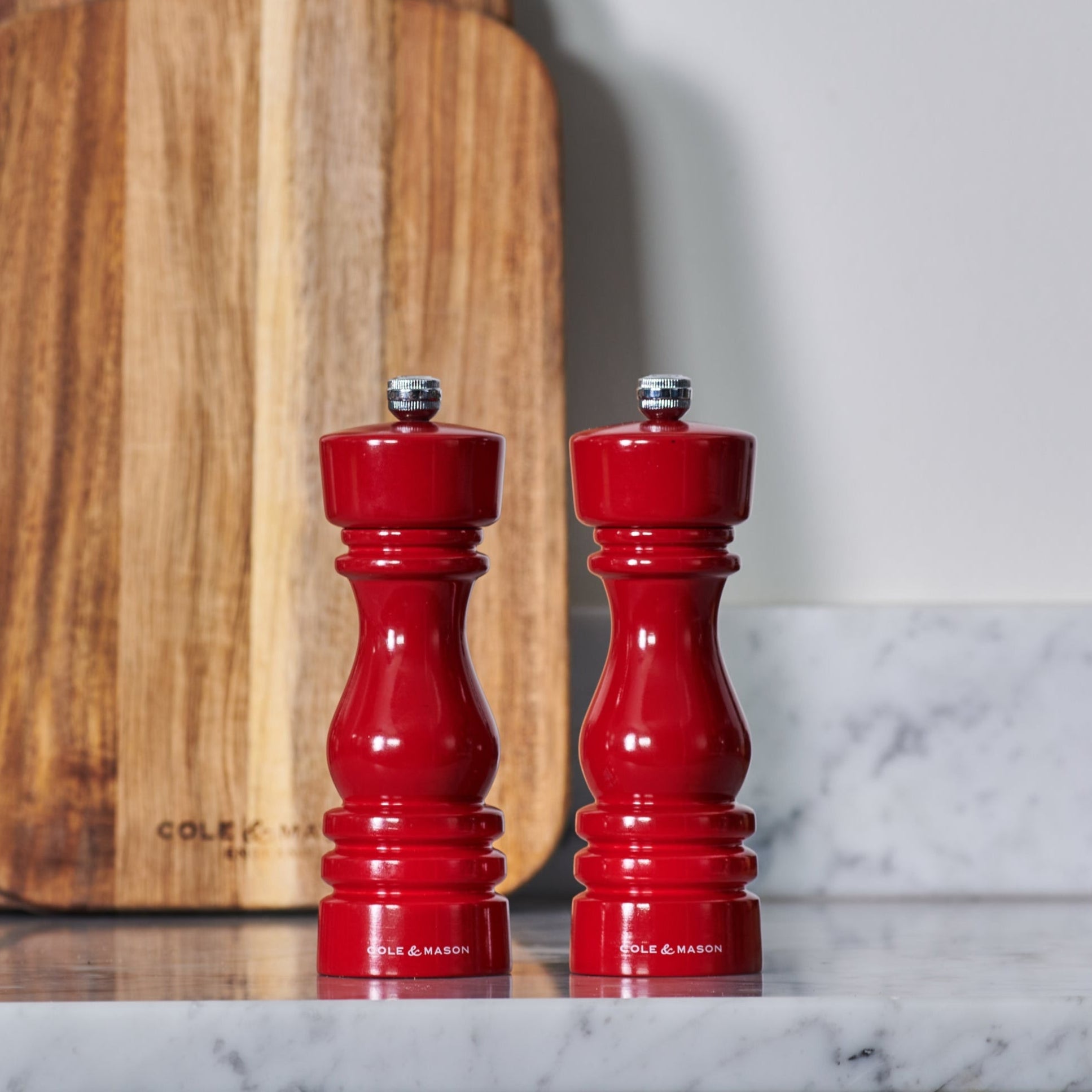 Two red salt and pepper mills sit on a white marble countertop in front of a wooden cutting board.