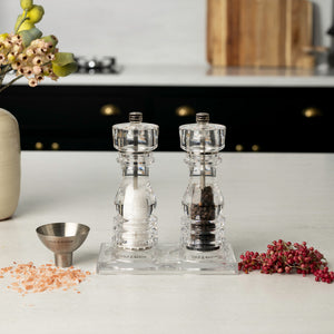 The Fast Bundle London Salt & Pepper Mill Set 180mm is displayed on a countertop tray with heaps of pink salt, black pepper, pink peppercorns, a small metal funnel, and flowers in a modern kitchen.