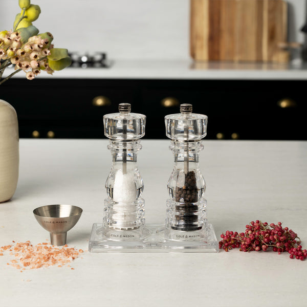 The Fast Bundle London Salt & Pepper Mill Set 180mm is displayed on a countertop tray with heaps of pink salt, black pepper, pink peppercorns, a small metal funnel, and flowers in a modern kitchen.