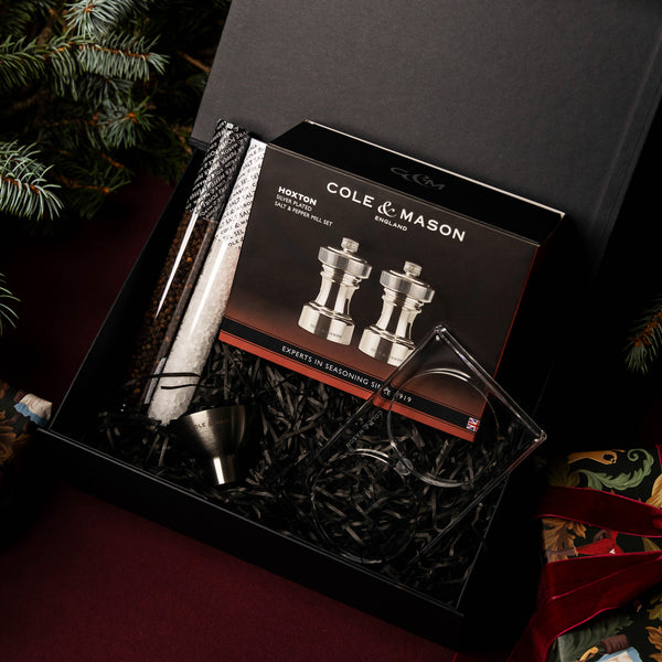 The Hoxton Silver Plated Mill Christmas Gift Hamper by Cole & Mason includes a salt and pepper mill set, metal funnel, two glass tubes with salt and pepper, and an acrylic stand on black shred—ideal for festive Christmas hampers.