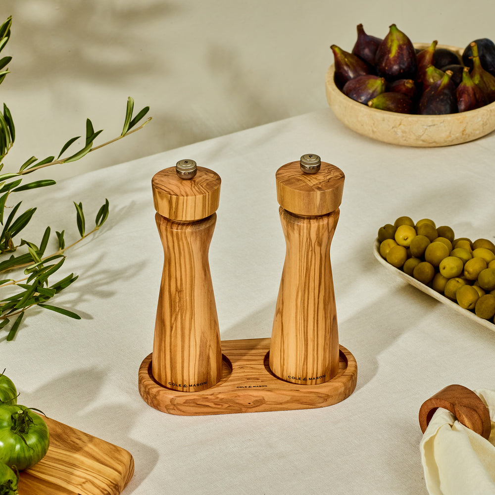 The Cole & Mason Portobello Set Bundle with M Board features wooden salt and pepper mills, an Italian olive wood board, green olives, chopped herbs, and a lidded salt cellar on a cream tablecloth with olive branches in the background.