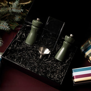 The Cole & Mason Kenton Mills in Rosemary Christmas Gift Hamper includes two green grinders, a metal funnel, and a double-sided jigger on black shredded paper, with pine branches and festive wrapped gifts for a cheerful holiday presentation.