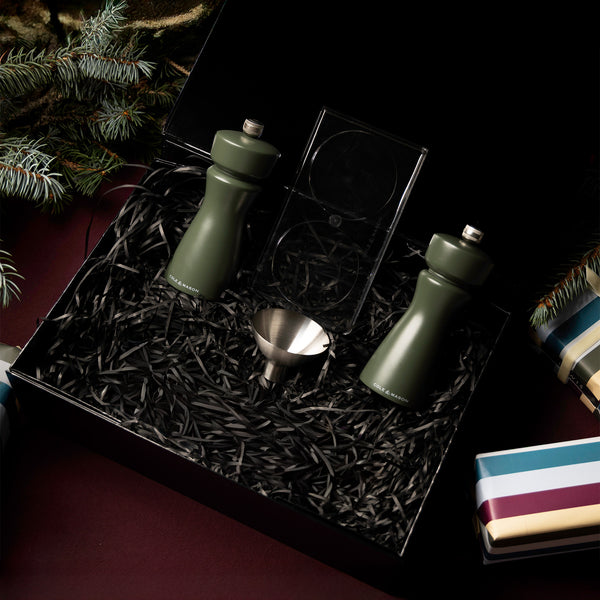 The Cole & Mason Kenton Mills in Rosemary Christmas Gift Hamper includes two green grinders, a metal funnel, and a double-sided jigger on black shredded paper, with pine branches and festive wrapped gifts for a cheerful holiday presentation.