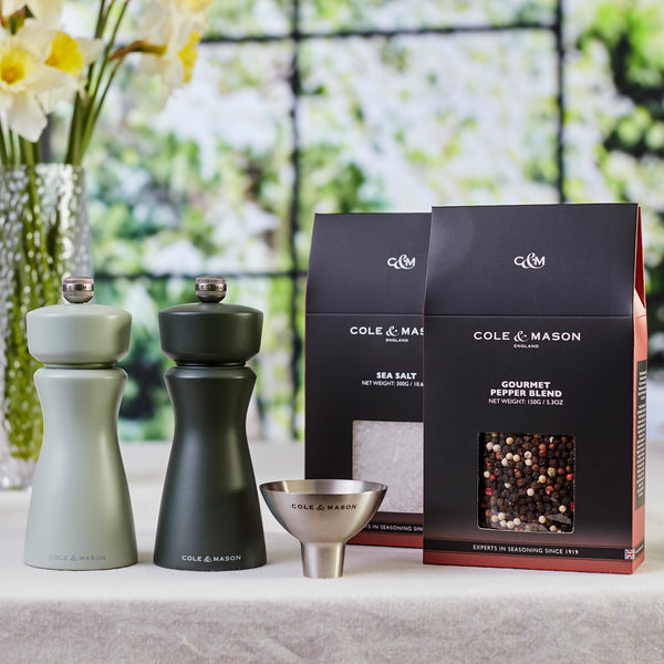Fast Bundle Mix & Match Kenton Mills salt and pepper grinders with ceramic mills, adjustable grind settings, a metal funnel, sea salt, and gourmet pepper blend are displayed on a kitchen counter with flowers in the background.