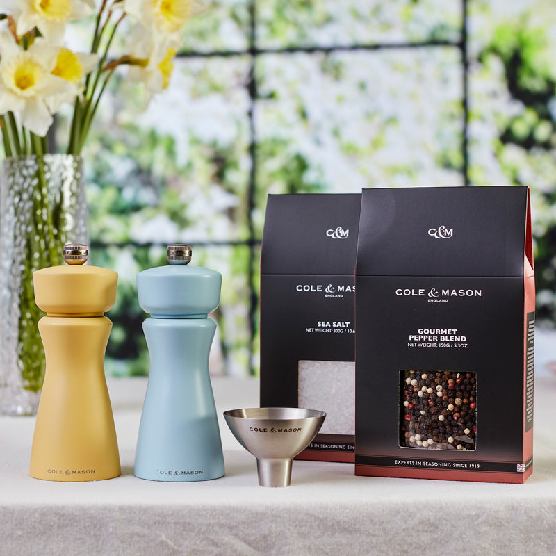 Two pastel-colored Mix & Match Kenton Mills salt and pepper grinders by Fast Bundle (yellow and blue), with adjustable grind settings, a metal funnel, and two boxed spice blends are displayed on a table with flowers by a sunny window.