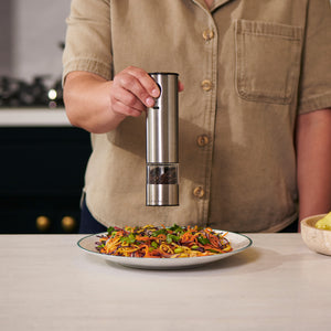 A person seasons a plate of colorful salad or noodles on the kitchen counter using the Cole & Mason Battersea USB Electric Salt & Pepper Mills with convenient one-handed operation.