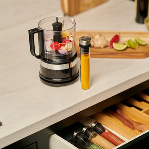 A food processor, yellow spice tube, and a cutting board with lime, ginger, and apple slices rest on a counter above the Cole & Mason Shadwell Drawer-Fit Spice Storage.