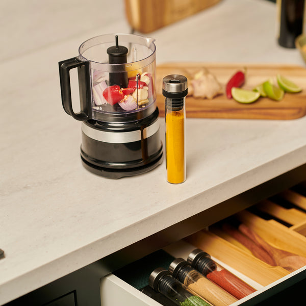 A food processor, yellow spice tube, and a cutting board with lime, ginger, and apple slices rest on a counter above the Cole & Mason Shadwell Drawer-Fit Spice Storage.
