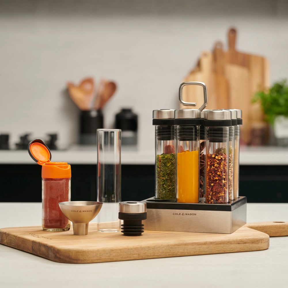 The Cole & Mason Shadwell Countertop Spice Storage features eight airtight glass jars in a stainless steel square holder with a metal handle for easy countertop organization.
