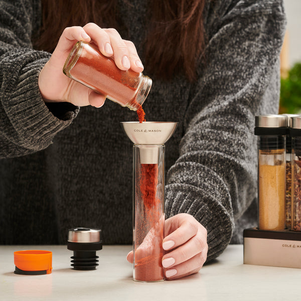 A person uses a metal funnel to transfer ground spice from a glass jar into the Shadwell Countertop Spice Storage by Cole & Mason, organizing spices with other neatly arranged jars on the counter.