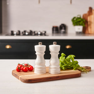 #salt&pepperset Cole & Mason’s London White Acrylic Salt & Pepper Mills 150mm, featuring an adjustable grinder, rest on a wooden cutting board with fresh tomatoes and greens, set on a kitchen counter with blurred cookware and stove in the background.
