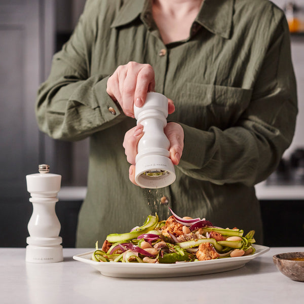 #salt&pepperset A person in a green shirt seasons salad using the Cole & Mason London White Acrylic Salt & Pepper Mill 150mm with adjustable grinding, while another high-gloss mill rests on the white countertop.