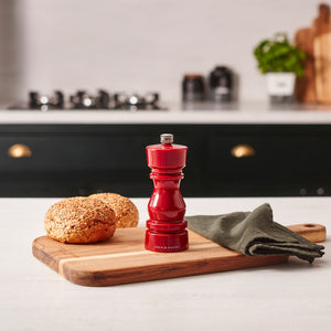 #pepper The Cole & Mason London Red Acrylic Salt & Pepper Mills 150mm are displayed on a wooden cutting board beside bread rolls and a green napkin, with a blurred kitchen stove and potted plant in the background.