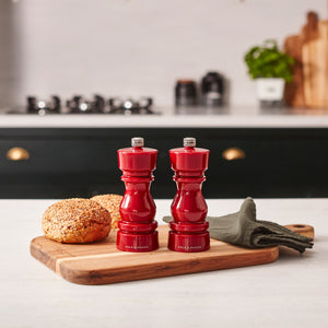 Two Cole & Mason London Red Acrylic Salt & Pepper Mills 150mm sit on a wooden board beside seeded bread rolls and a dark folded napkin, with a stove and potted plant softly blurred in the background.