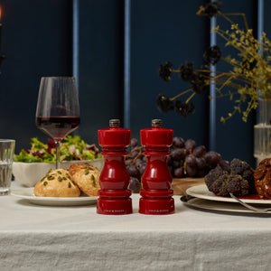 #pepper A table set with Cole & Mason London Red Acrylic Salt & Pepper Mills 150mm with adjustable grinding, a glass of red wine, salad, bread rolls, grapes, and a plate of food against a dark blue wall and flowers.