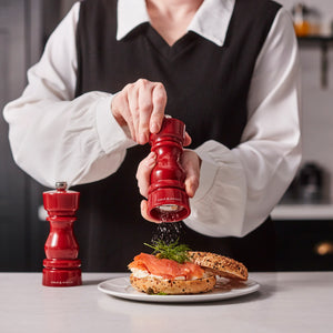 #salt&pepperset A person in a white blouse and black vest uses a Cole & Mason London Red Acrylic Salt & Pepper Mill 150mm to grind pepper onto a bagel with salmon and dill. Another matching mill sits on the table beside the plate.