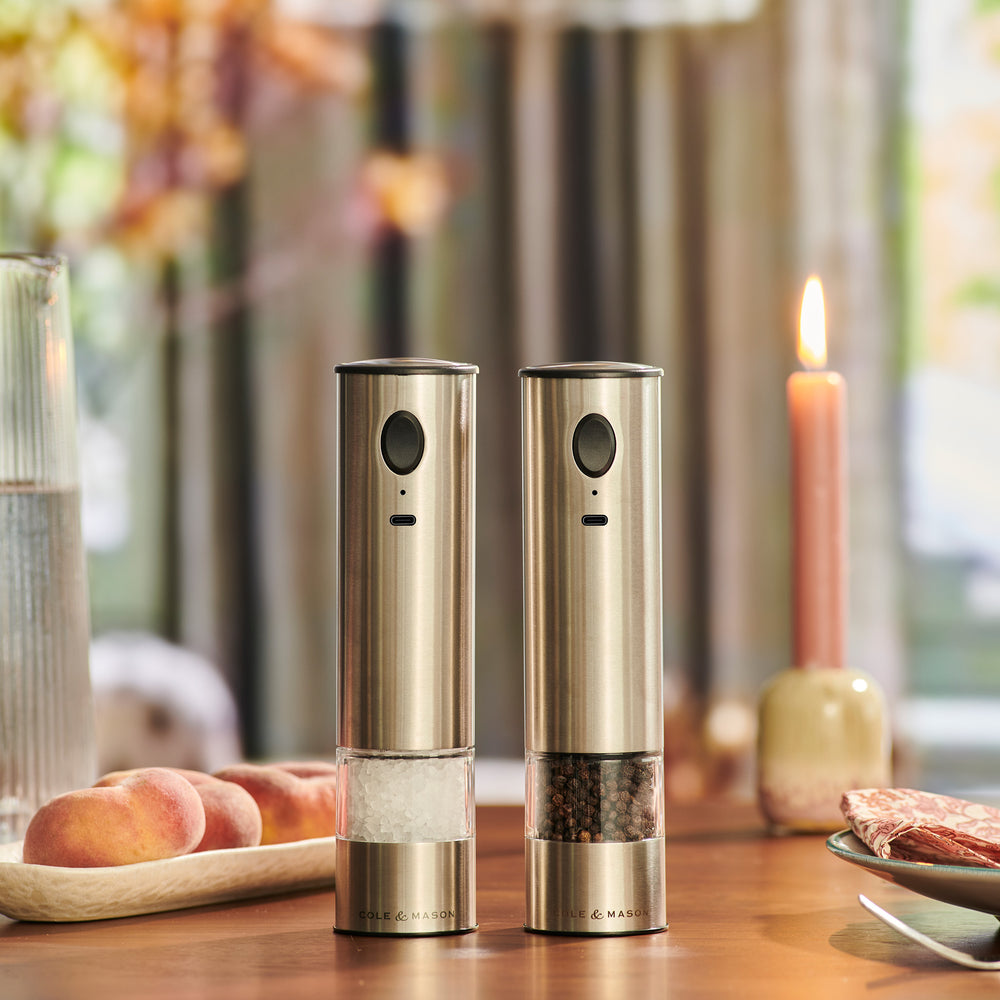 The Cole & Mason Battersea USB Electric Salt & Pepper Mill features a stainless steel design, ceramic grinder, transparent chamber, and front button for one-handed use. It stands upright against a light background.