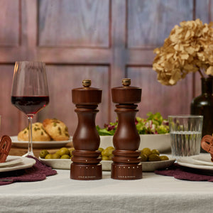 A set dining table features two wooden grinders, including the elegant Cole & Mason London Walnut Salt or Pepper Mill, accompanied by a glass of red wine, salad, olives, bread, plates, and dried flowers in the background.