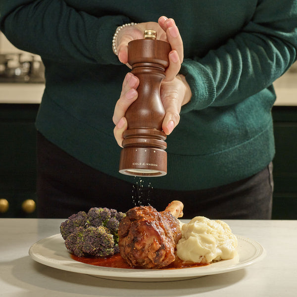 A person uses the Cole & Mason London Walnut Salt or Pepper Mill to grind pepper onto a plate with mashed potatoes, broccoli, and a meat dish in red sauce.