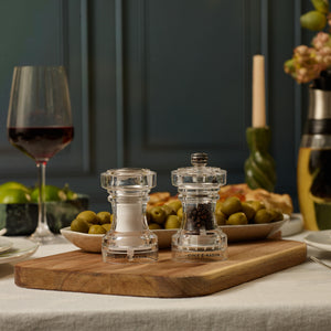 Two Cole & Mason Acrylic Salt Shaker & Pepper Mill Set, 104 mm, with adjustable grinder settings rest on a wooden board amidst bowls of green olives, a glass of red wine, and a partially visible candle, all arranged on a white tablecloth.