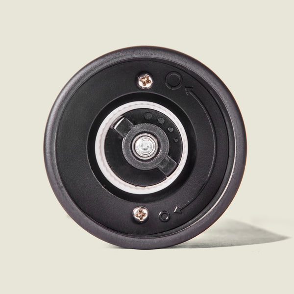 Close-up of the bottom of the Cole & Mason Brixton Electric Mill Matt Black GS, showcasing its ceramic grinder and adjustable grind mechanism with metal screws and embossed directional arrows for grind size adjustment—all thoughtfully designed for easy one-handed operation, set against a plain light background.