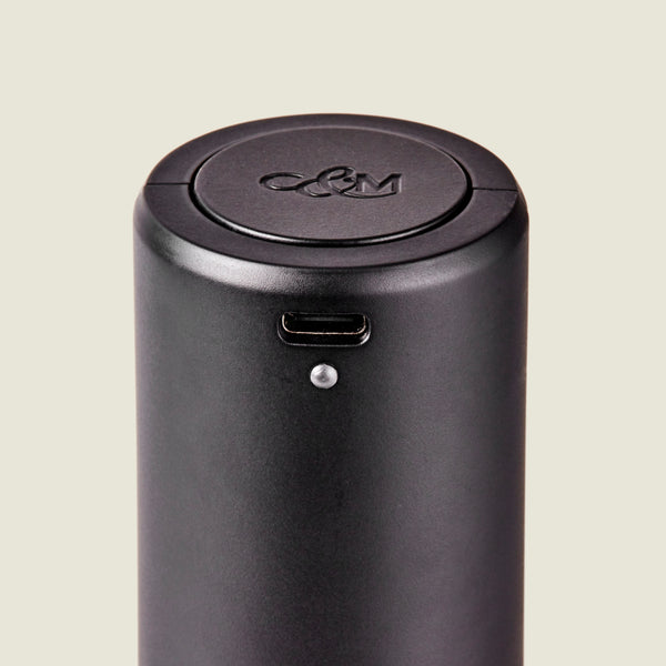 Close-up of the Cole & Mason Brixton Electric Mill Matt Black GS, a cylindrical black electronic device featuring a USB-C port and a small indicator light. Designed for one-handed operation, this electric mill has an embossed C&M logo on its circular lid and contains a durable ceramic grinder inside.