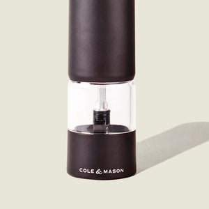 A black and clear Cole & Mason Brixton Electric Mill Matt Black GS pepper grinder with a ceramic mechanism stands upright on a light background, casting a faint shadow to the right.