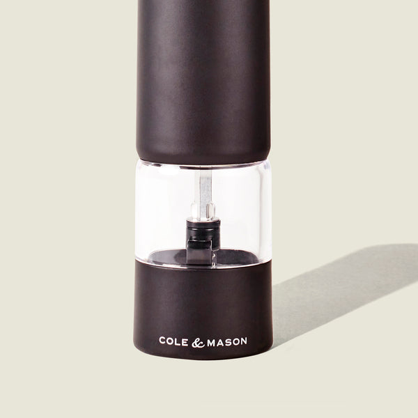 A black and clear Cole & Mason Brixton Electric Mill Matt Black GS pepper grinder with a ceramic mechanism stands upright on a light background, casting a faint shadow to the right.