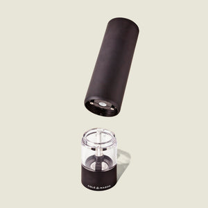 The Cole & Mason Brixton Electric Mill Matt Black GS is pictured in two separate parts on a light beige background. The top cylindrical black section sits above the transparent base, revealing the ceramic grinding mechanism inside and showcasing its convenient one-hand operation.