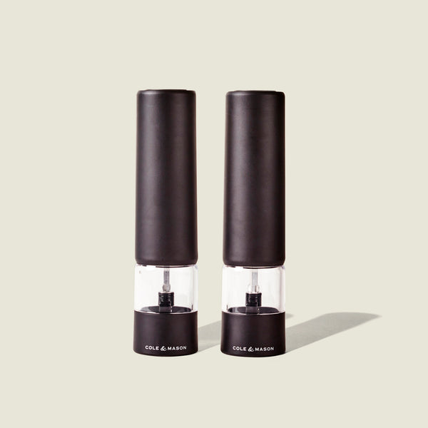 Two sleek, matt black electric grinders labeled Cole & Mason Brixton Electric Mill stand side by side against a plain, light background, each featuring a durable ceramic grinder for smooth and precise seasoning with convenient one-hand operation.
