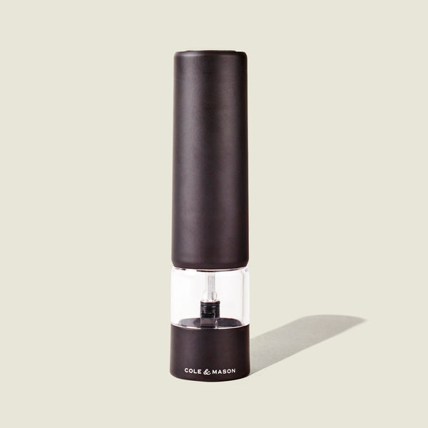 The Cole & Mason Brixton Electric Mill Matt Black GS is an electric pepper grinder with a ceramic grinding mechanism and a clear middle section for easy viewing, designed for convenient one-hand operation. The mill, branded with the Cole & Mason logo on its base, stands upright against a light beige background.