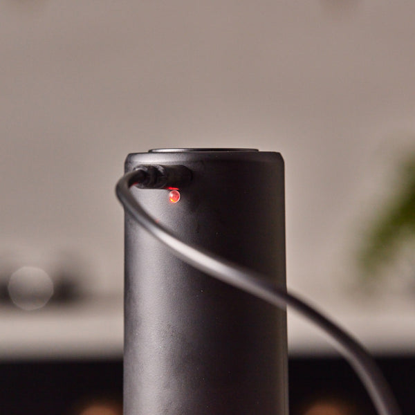 A Cole & Mason Brixton Electric Mill Matt Black GS, featuring a sleek cylindrical design with a power cord plugged in and a small red indicator light glowing near the port, is showcased against a blurred background that accentuates its convenient one-hand operation in the foreground.