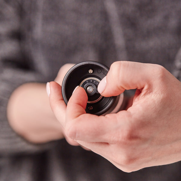 A person uses one hand to adjust the settings on the Cole & Mason Brixton Electric Mill Matt Black GS, a small, round electronic device, with their thumb and index finger. The background is out of focus.