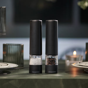 Two Brixton Electric Mill Matt Black GS grinders by Cole & Mason, featuring convenient one-hand operation and a modern design, sit side by side on a green table, with plates, glassware, and a lit candle softly blurred in the background.